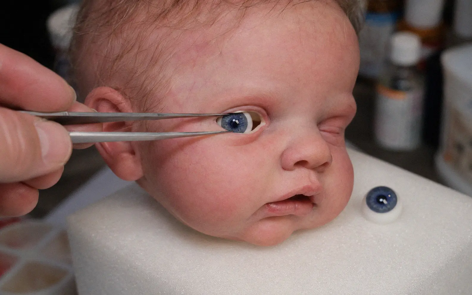 Tweezers positioning a glass eye into the eye socket of a silicone baby doll head