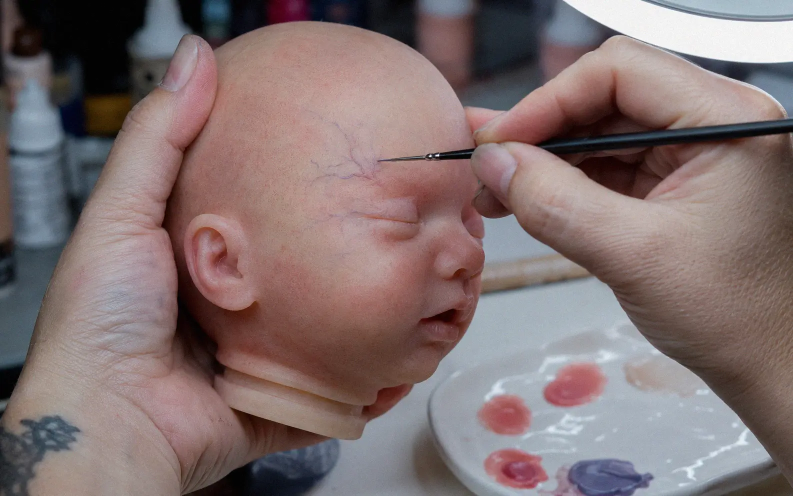 Artist applying thin layers of silicone paint to a silicone baby doll head with a fine brush