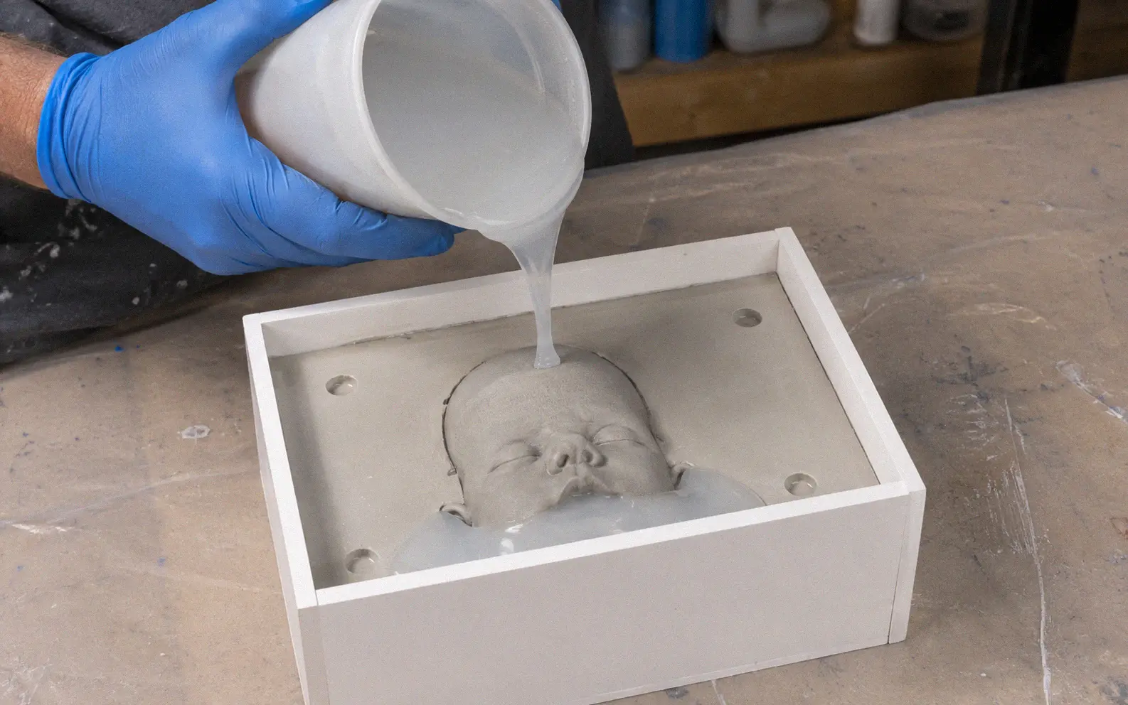 Silicone rubber being poured into a LEGO mold box over a clay baby head sculpture