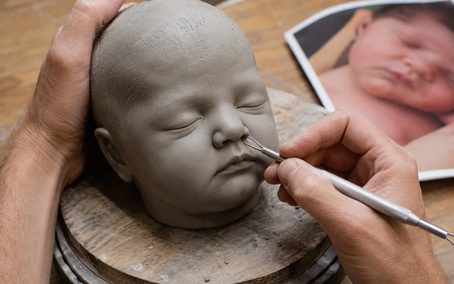 Artist's hands sculpting a realistic baby face from gray clay on a wooden turntable